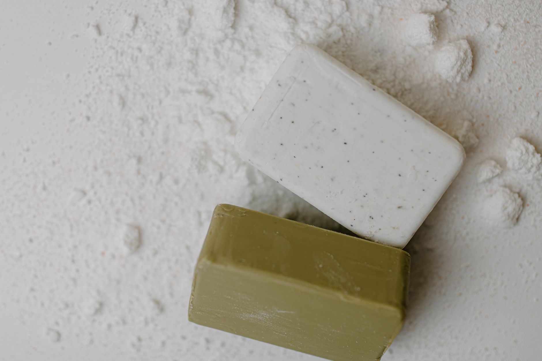 Deux pains de savon opaques saponifiés à froid, l'un blanc exfoliant, l'autre vert olive, posés sur une base poudreuse